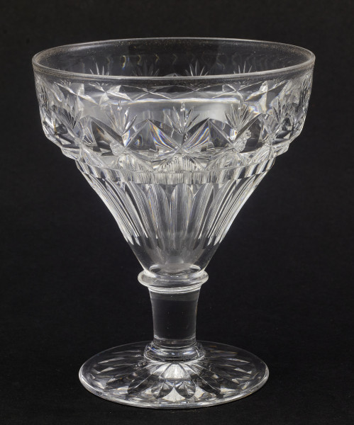 Stopa okrągła, nóżka krótka i szeroka, zakończona pierścieniem, na którym osadzona stożkowata czara. Spód stopy dekorowany płytkim szlifem o motywie wielopłatkowego kwiatka.