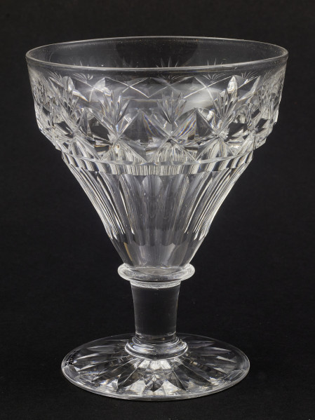 Kieliszek duży, wykonano ze szkła kryształowego
Stopa okrągła, nóżka krótka i szeroka, zakończona pierścieniem, na którym osadzona stożkowata czara. Spód stopy dekorowany płytkim szlifem o motywie wielopłatkowego kwiatka.
