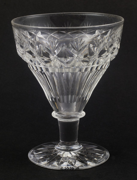 Kieliszek duży, wykonano ze szkła kryształowego
Stopa okrągła, nóżka krótka i szeroka, zakończona pierścieniem, na którym osadzona stożkowata czara. Spód stopy dekorowany płytkim szlifem o motywie wielopłatkowego kwiatka.
