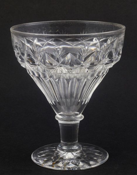 Stopa okrągła, nóżka krótka i szeroka, zakończona pierścieniem, na którym osadzona stożkowata czara. Spód stopy dekorowany płytkim szlifem o motywie wielopłatkowego kwiatka.