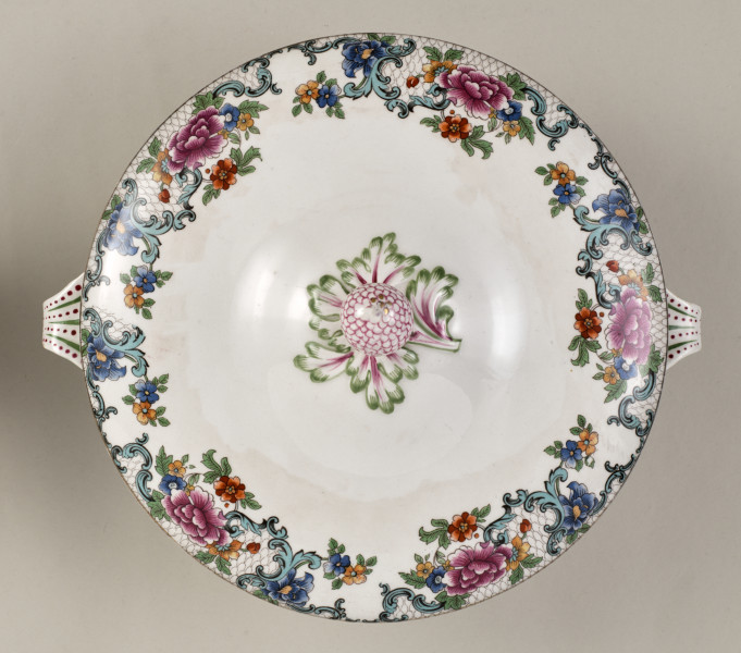 Półmiskowa waza z pokrywką, wykonana z białego fajansu, obiekt w kształcie cylindra. Naczynie ozdobione wielobarwną kalkomanią złożoną z asymetrycznych roślinnych kompozycji, wzory nawiązują do sztuki rokoko.