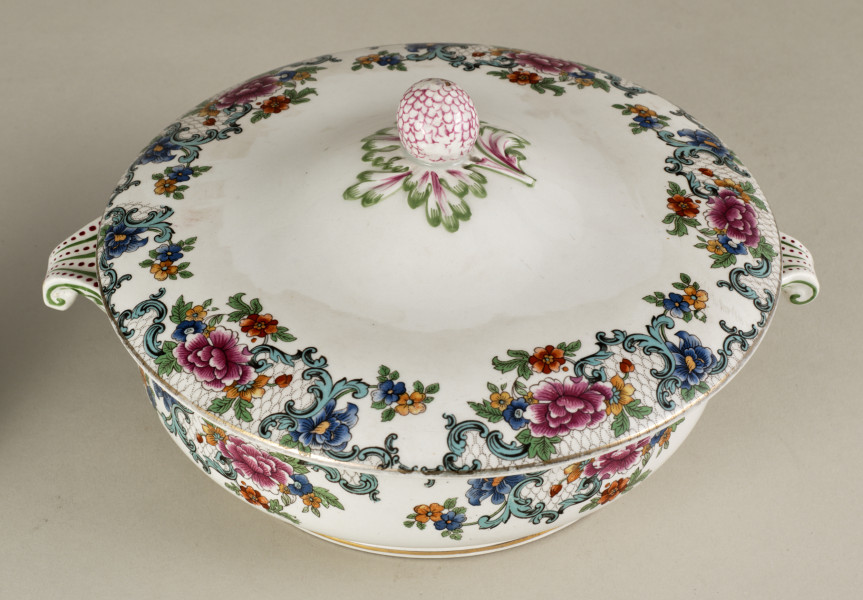 Półmiskowa waza z pokrywką, wykonana z białego fajansu, obiekt w kształcie cylindra. Naczynie ozdobione wielobarwną kalkomanią złożoną z asymetrycznych roślinnych kompozycji, wzory nawiązują do sztuki rokoko.